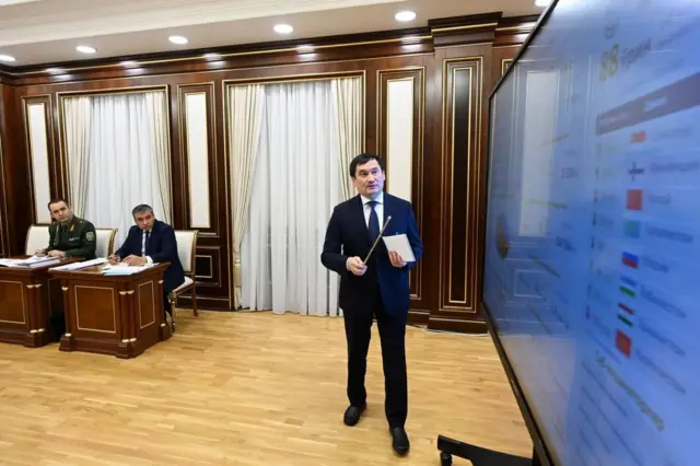 Rasmiy eraka qora kostyum shimda ukaska ushlagan holda xonaga o'tirgnalarga oq taxtaga qarab tushuntiryapti, taxtada bayroqlari bilan davlatlar nomlanishi yozilgan