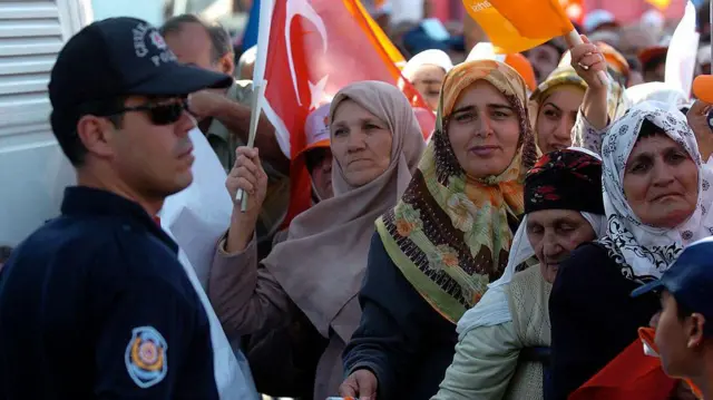 Bir grup türbanlı kadın, ellerinde Türk bayrağı ve AKP bayrakları var. Önlerinde bir erkek polis duruyor. 