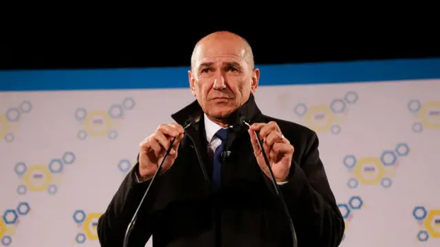 Janez Janša u crnom kaputu za govornicom, rukama drži dva mikrofona