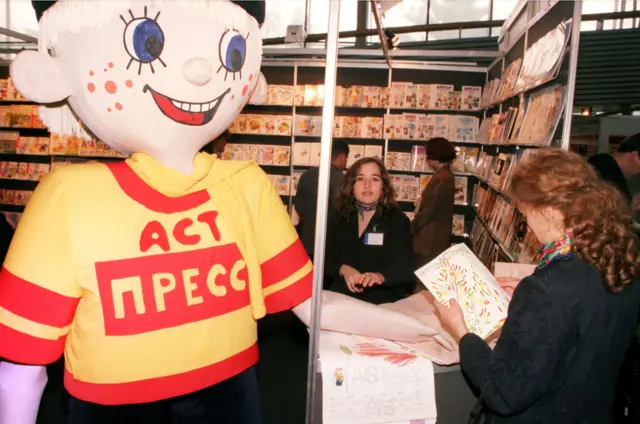 Ростовая кукла в желтой в футболке с красной надписью «АСТ Пресс». На заднем плане — стеллажи с книгами и журналами и посетители. 