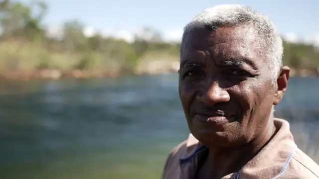 Homem negro com cabelos brancos, vestido com camisa polo, posa para a câmera com rio de águas azuladas ao fundo