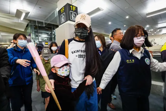 大陆配偶网红“亚亚”在台湾的机场，她戴上口罩和米黄色的鸭舌帽，拥着一名儿童，旁边有身穿写有“移民署”的人士陪同。