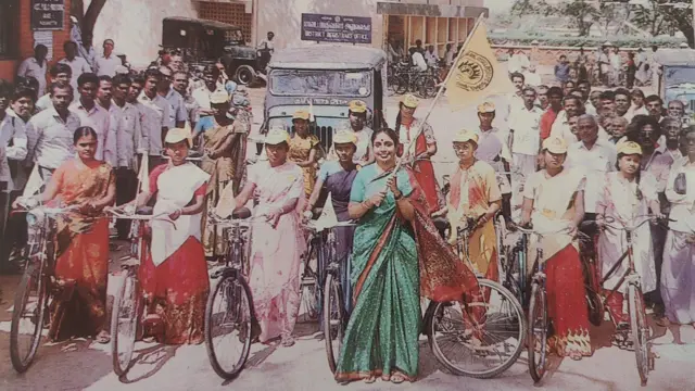 Sheela Rani Chunkath vestindo um sári verde, de pé, em frente a meia dúzia de ciclistas vestindo meios-sáris