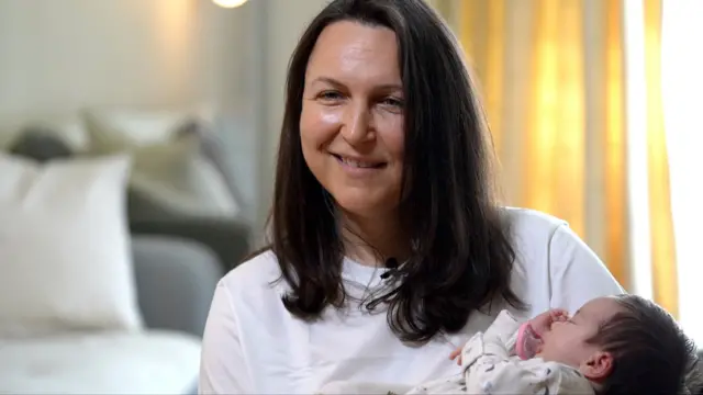 Ана держит на руках своего ребенка, она в белой футболке, у нее длинные каштановые волосы