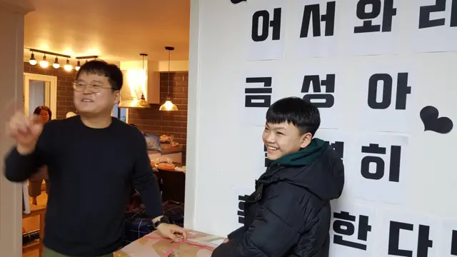 一张照片中，一个笑容满面的青少年和他的寄养父亲站在一起，身边有箱子，背景是温馨的家，他身后的文字是韩文写的“欢迎回家，金城！”