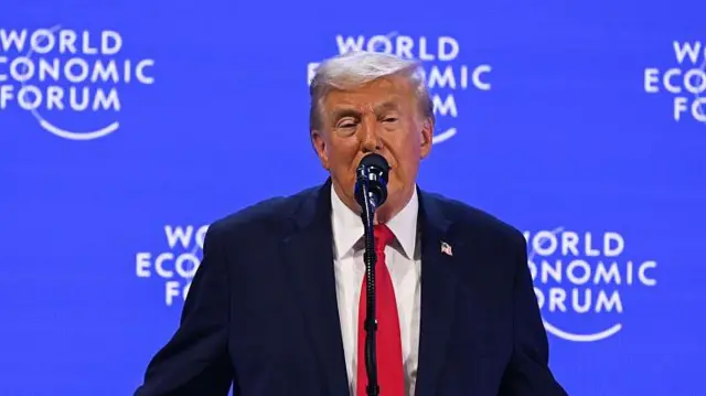 Kırmızı kravatlı lacivert takım elbiseli adam kürsüde konuşuyor. Arkasındaki mavi ekranda World Economic Forum yazıyor. 
