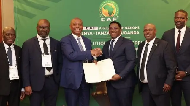 Un groupe de quatre ministres des Sports et dirigeants du football originaires de Tanzanie et d’Ouganda, tous vêtus de costumes sombres, de chemises blanches et de cravates foncées, posent pour une photo aux côtés du président de la Confédération africaine de football, Patrice Motsepe (troisième à partir de la gauche), et du secrétaire général Samson Adamu (tout à droite), habillés de manière similaire. Le groupe se tient devant un panneau vert portant le logo de la CAF, et Motsepe serre la main de l’un des responsables tandis que les deux hommes tiennent un grand document ouvert face à la caméra.