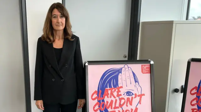 Alison está de pie en el interior junto a un gran póster en un caballete. El póster presenta un texto en negrita en rojo y azul que dice "CLARE NO PUDO, PERO TÚ SÍ. TIENES DERECHO A PREGUNTAR", junto con texto y gráficos más pequeños. La persona viste un traje negro y tacones, y el fondo incluye una puerta y un armario.