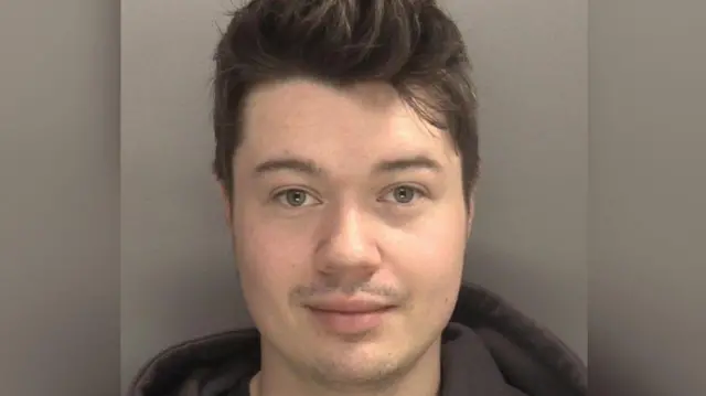 A mugshot showing the face of a young man with short dark hair and a dark hoodie against a grey background.