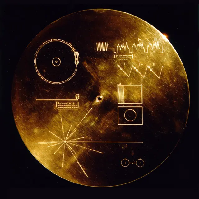 A photo of a circular golden disc engraved with several symbols and drawings, such as a zigzag wave pattern, rectangles and circles, and a point with many lines emanating from it.