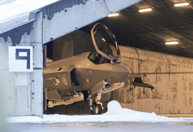 Parte delantera de un caza F-35 visto en el interior de un hangar. Hay nieve en el suelo y parece haberla también sobre el techo del hangar. 