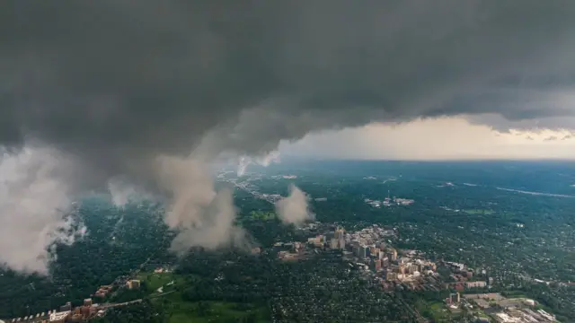 Vista aérea de uma cidade com abundantes áreas verdes e grupos de edifícios dispersos, coberta por nuvens escuras