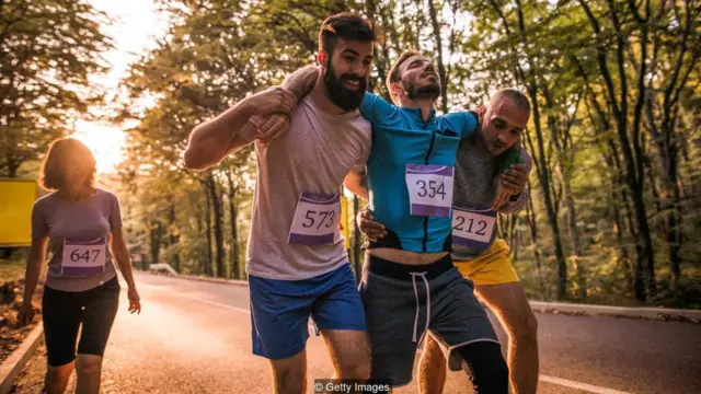 Dois maratonistas carregam um terceiro entre eles, machucado; os três homens são observados por uma outra corredora, mulher