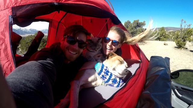 O casal Cynthia e Andre posa para uma selfie junto com o gato e o cachorro, dentro de uma cabana no meio das montanhas