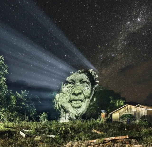 A man's face is projected onto the Brazilian rainforest canopy