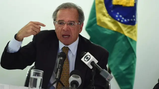 Paulo Guedes sentado à mesa falando com diversos microfones a frente