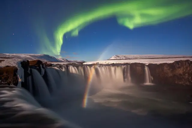 A aurora boreal e um arco-íris lunar visto acima da cachoeira Godafoss, na Islândia