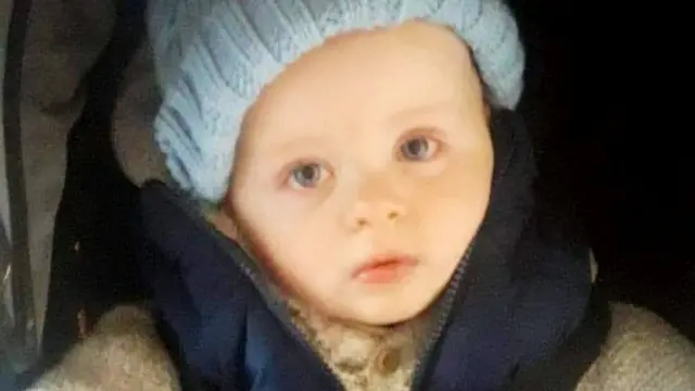 Um bebê branco de olhos azuis sentado em uma cadeirinha de carro