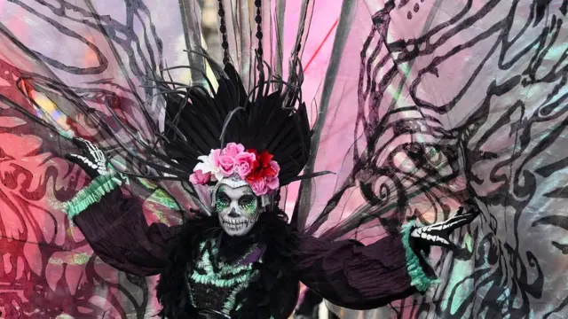 Человек в черном костюме с венком из малиновых цветов, прозрачными крыльями, и лицом, раскрашенным под череп