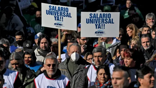 Várias pessoas protestando na rua, duas delas segurando cartaz dizendo: 'Salário universal ya!"