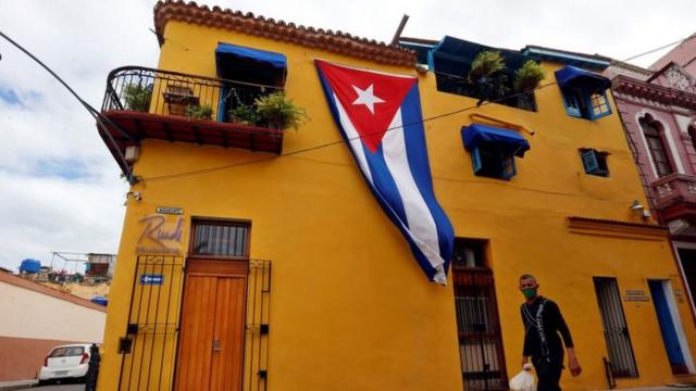 Homem caminharoleta de parque de diversãoruaroleta de parque de diversãoCuba. Acima dele aparece uma bandeira do país pendurada na fachadaroleta de parque de diversãouma casa.