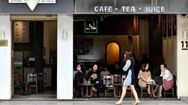 A woman wearing a mask walks past a cafe where customers sit and chat