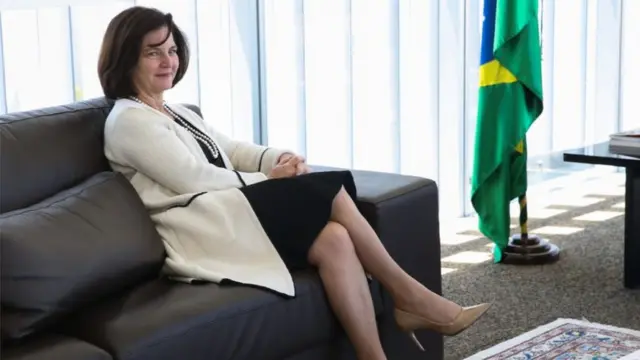 A procuradora Raquel Dodge sentadaapk vaidebetum sofá escuro ao ladoapk vaidebetuma bandeira do Brasil