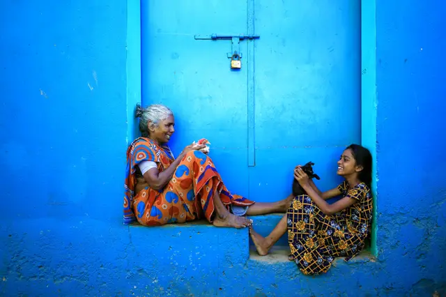 An elderly woman and a girl sit in a blue doorway with a black kitten and a chicken
