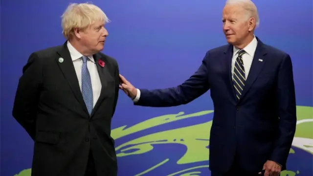 Johnson e Biden em pé, em frente a painel de evento, sorrindo timidamente ao conversarem