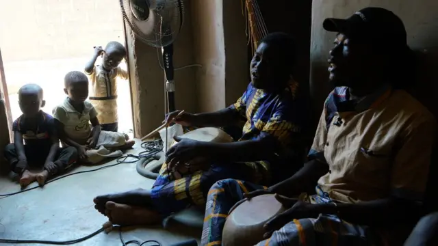 (De droite à gauche) Maboudou Sanou et Ibrahim Dembélé chantent pendant une répétition dans la maison que Sanou partage avec le compositeur Keiko Fujiie, une semaine avant la première de l'opéra Là-bas ou Ici (Here or There) à Ouagadougou. Le fils de Sanou et des enfants du quartier écoutent.