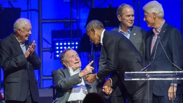 ජ්‍යෙෂ්ඨ බුෂ්, ඔබාමාගේ අතගෙන යමක් පවසද්දී කාටර් සතුටින් අත්පොලසන් නගයි. ක්ලින්ටන් සහ බුෂ් (කණිෂ්ඨ) කතාබහක.