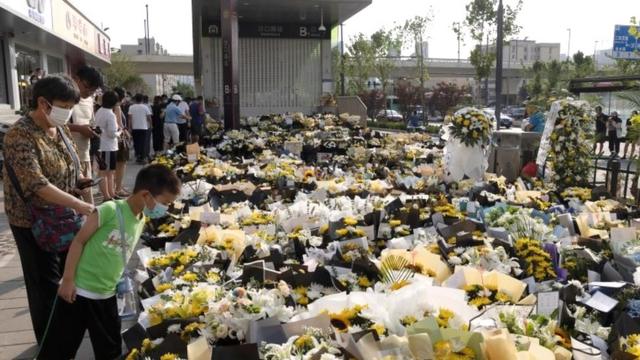 People stand next to flowers placed at an entrance to a subway station in Zhengzhou