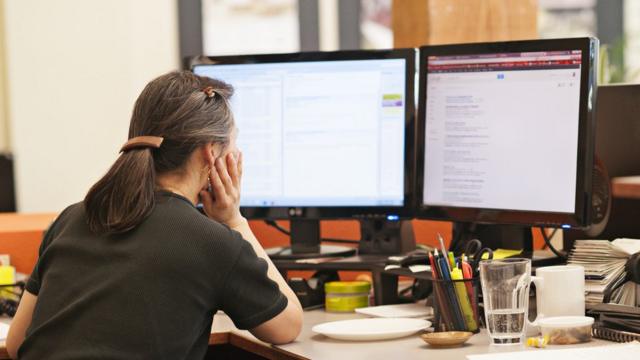 Mulher de cabelo grisalho, sentada e de costas olha para duas telas de computador em escritório