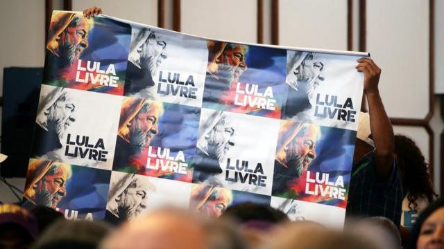 Em meio a plateia em evento, cartaz é levantado com diversas fotos de Lula e dizeres 'Lula Livre'