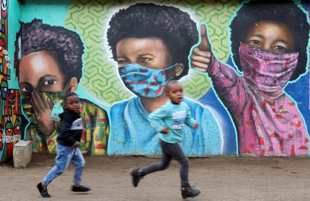Boys run past a bright mural of children wearing face masks.