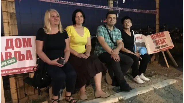 Četvero stanara sede noću ispred žičane ograde sa transparentima