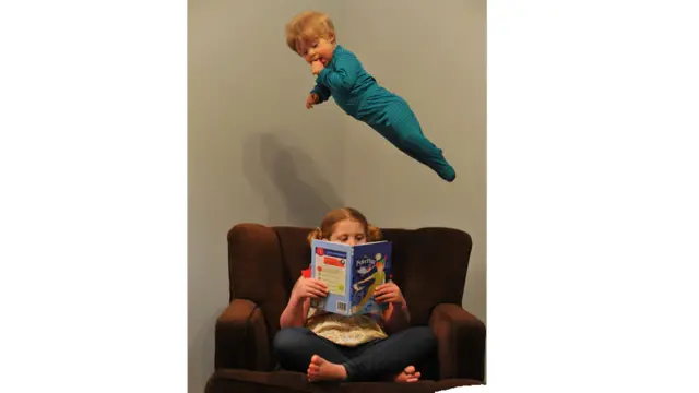 Will en el aire como si estuviera volando sobre su hermana sentada en un sillón leyendo un libro