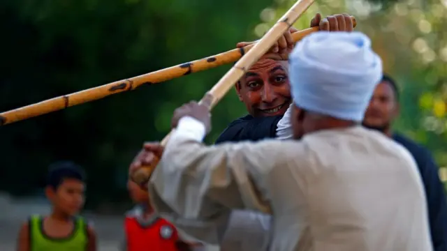رجلان يرقصان رقصة التحطيب وخلفهما طفلين ورجل يشاهدان الرقص