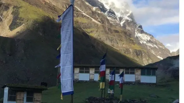 A single-story village home with peaked roof, located alongside a steep mountainside, with grass at the base and snow on rocky peaks
