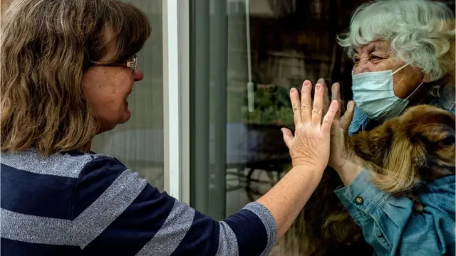 Duas mulheres separadas por um vidro se cumprimentam