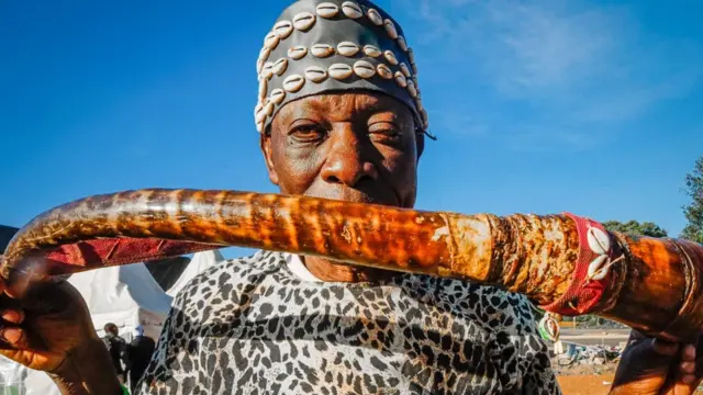 Mutum sanye da tufafin gargajiya, dauke da kahon da ya ke amfani da shi wajen kida a hannunsa.