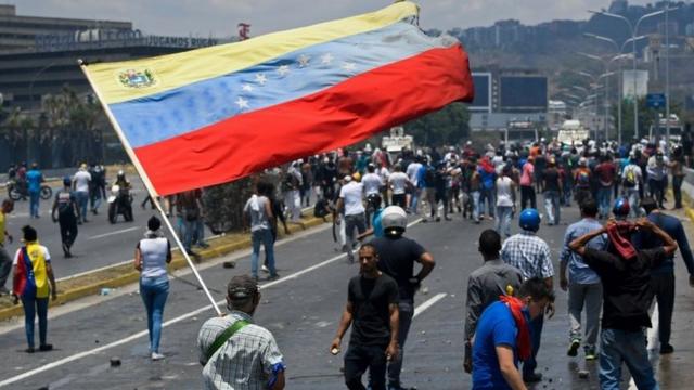 An opposition demonstrator holds a large Venezuelan flag amid a large scattered crowd