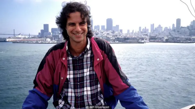 Jeff Greenwald, em foto antiga, sorrindo em margem de baía, com prédios ao fundo