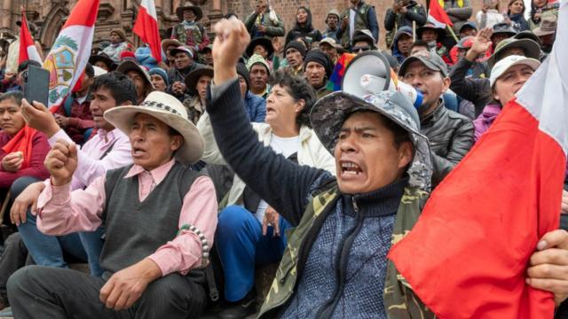 Manifestantes sentados gritam e seguram bandeiras do Peru