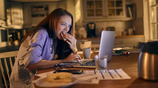 شابة تتناول الطعام وهي تحدق في شاشة جهاز كمبيوتر محمول