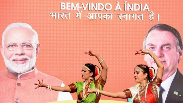 Duas dançarinas com trajes tradicionais fazem movimento em frente a painel com fotos de Narendra Modi e Jair Bolsonaro, além dos dizeres: 'Bem-vindo à Índia'