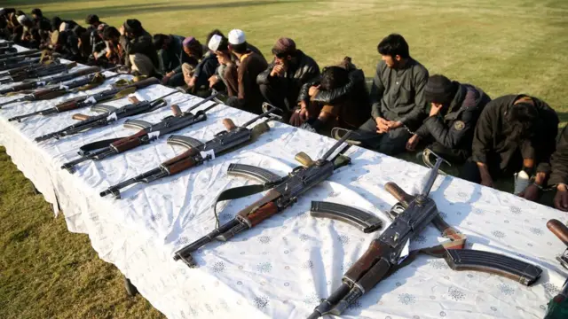 Membros do Talebã e Estado Islâmico escondemo os rostos diante de mesa com diversas armas enfileiradas