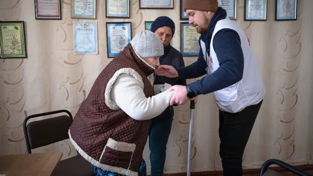 Maria parándose de una silla con la ayuda de un médico que la toma de las manos