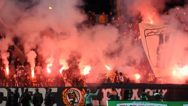Football supporters for Mhiri stadium, Tunisia dey wave flares and flags during friendly match against France. Dem dey celebrate dia club 90 years anniversary