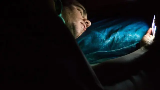 شاب يتصفح هاتفه المحمول وهو نائم على فراشه